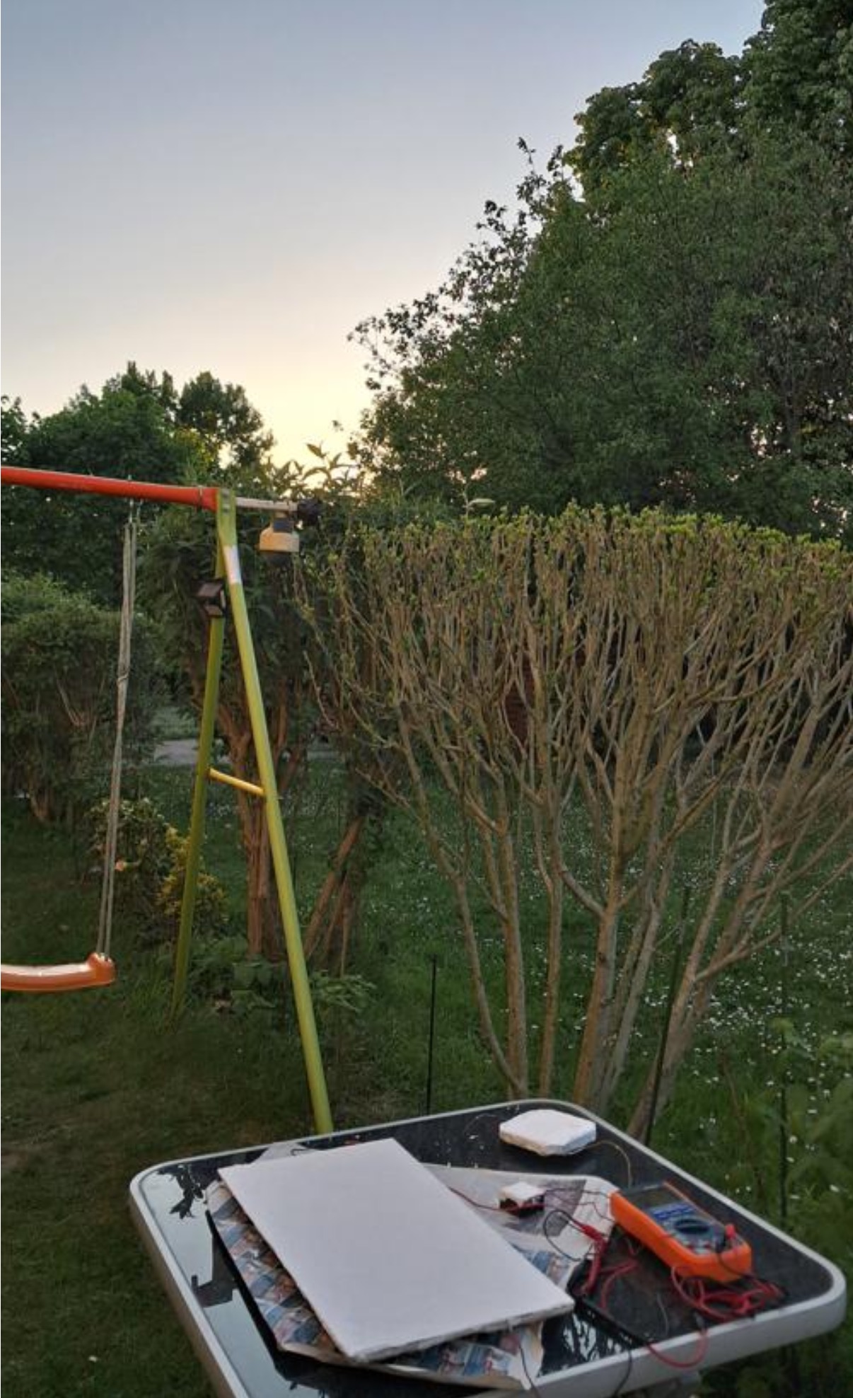 A white CryoPaint-coated panel laid on a garden table at dusk in Mannheim, Germany, wired to a multimeter for a radiative-cooling thermoelectric generator test.